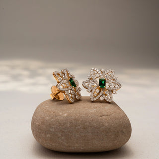 Angled view of floral diamond stud earrings in 14 KT Yellow Gold, featuring pavé-set looping petals with emerald-cut green center stones, displayed on a textured stone surface.
