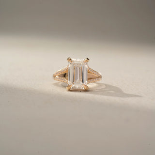 Front view of an emerald-cut engagement ring in 14 KT Rose Gold, featuring an emerald-cut lab-grown diamond with a pavé-set split shank band, displayed against a beige backdrop.