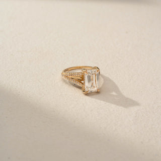 Side view of an emerald-cut engagement ring in 14 KT Rose Gold, featuring an emerald-cut lab-grown diamond with a pavé-set split shank band, displayed against a beige backdrop.
