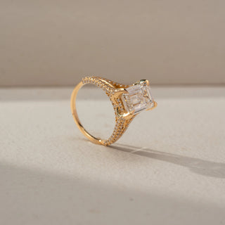 Angled view of an emerald-cut engagement ring in 14 KT Rose Gold, featuring an emerald-cut lab-grown diamond with a pavé-set split shank band, displayed against a beige backdrop.