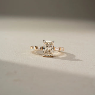 Front view of a yellow gold ring featuring a radiant-cut center diamond with tapered baguette side diamonds, displayed on a soft ivory surface under warm natural light.