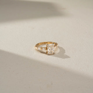 Top view of a yellow gold ring featuring a radiant-cut center diamond with baguette-cut side diamonds along the band, displayed on a soft ivory surface in warm natural light.