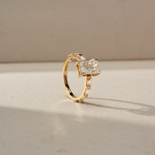 Angled view of a yellow gold ring featuring a radiant-cut center diamond with baguette-cut diamond accents on the band, showcased on a neutral surface in soft natural light.