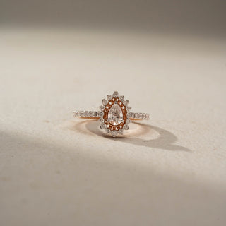 Front view of a pear halo engagement ring in 14 KT Rose Gold, featuring a pear-shaped lab-grown diamond framed by a rose gold halo and surrounded by a scalloped diamond halo, displayed against a beige backdrop.
