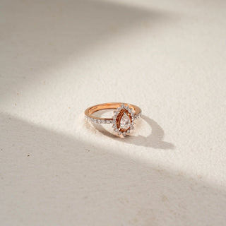 Side view of a pear halo engagement ring in 14 KT Rose Gold, featuring a pear-shaped lab-grown diamond framed by a rose gold halo and surrounded by a scalloped diamond halo, displayed against a beige backdrop.