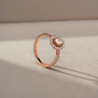 Angled view of a pear halo engagement ring in 14 KT Rose Gold, featuring a pear-shaped lab-grown diamond framed by a rose gold halo and surrounded by a scalloped diamond halo, displayed against a beige backdrop.