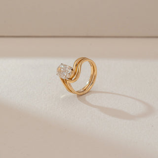 Side profile of oval diamond ring in yellow gold, featuring a central lab-grown diamond with a flowing double wave band. Captured against a soft beige textured background with gentle shadows.