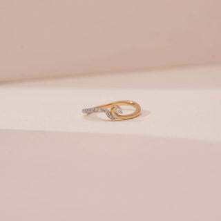 Front view of Reia’s 14 KT Yellow Gold Modern Swirl Diamond Band. The intricate bypass design and shimmering pavé-set details are showcased against a soft pink surface with gentle shadows.