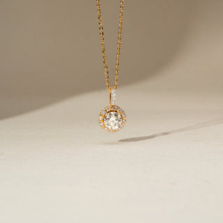 Front view of a gold pendant necklace featuring a round solitaire diamond surrounded by a halo of smaller diamonds, suspended from a delicate chain against a soft cream background.