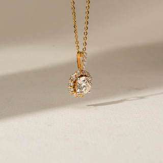 Side view of a gold halo pendant necklace featuring a round center diamond surrounded by smaller diamonds, showcasing the raised setting and diamond-accented bail on a cream background.