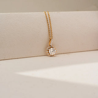 Close-up view of a gold halo pendant necklace featuring a brilliant round center diamond surrounded by smaller diamonds, suspended from a delicate chain against a soft cream backdrop.