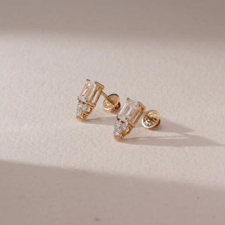 Side view of diamond stud earrings in 14KT yellow gold, featuring emerald-cut diamonds with marquise side accents and screw backs, set on a beige surface highlighting intricate design and brilliance.
