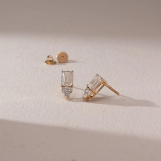 Angled view of diamond stud earrings in 14KT yellow gold, featuring emerald-cut diamonds with marquise accents and screw backs, placed on a beige surface highlighting fine detail and brilliance.
