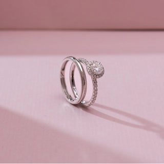 Angled view of Timeless Pavé Accented Diamond Ring in white gold, featuring a round lab-grown diamond halo with pavé-set band, displayed stacked with matching bands on a soft pink backdrop.