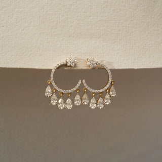 Front view of a pair of diamond hoop earrings with floral stud tops, pavé-set hoops, and dangling pear-shaped diamond drops, displayed on a textured cream-colored surface.