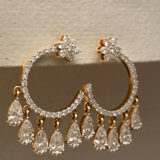 Close-up view of diamond hoop earrings with floral cluster tops, pavé-set hoops, and multiple dangling pear-shaped diamonds, displayed on a textured cream-colored surface.