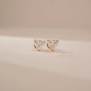 Front view of heart-shaped diamond stud earrings in 14KT Yellow Gold, featuring prong settings on a beige backdrop.