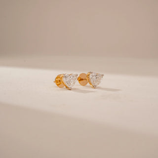 Side view of heart-shaped diamond stud earrings in 14KT Yellow Gold, featuring prong settings on a beige backdrop.