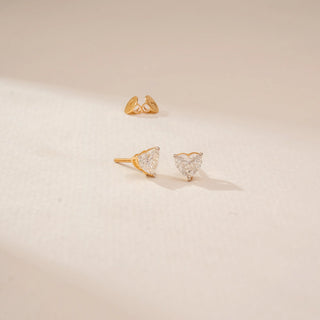 Angled view of heart-shaped diamond stud earrings in 14KT Yellow Gold, featuring prong settings on a beige backdrop.