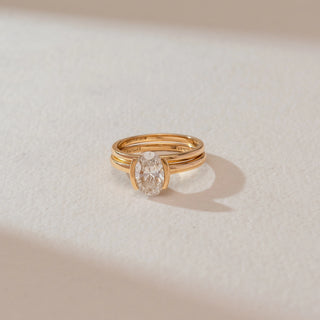 Top angled view of oval diamond ring in yellow gold, featuring a central lab-grown diamond held between parallel band accents. Set against a soft beige textured background with natural shadows.