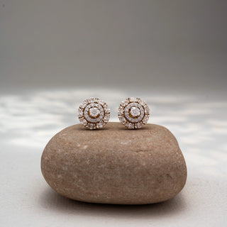 Front view of round diamond stud earrings in 14 KT Yellow Gold, featuring concentric halo circles with brilliant center diamonds, displayed on a textured stone surface.
