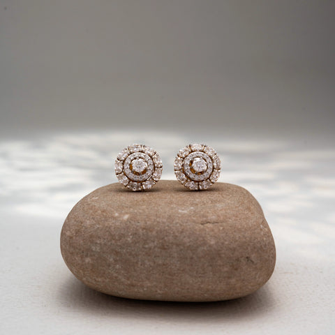 Front view of round diamond stud earrings in 14 KT Yellow Gold, featuring concentric halo circles with brilliant center diamonds, displayed on a textured stone surface.
