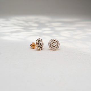Angled view of round diamond stud earrings in 14 KT Yellow Gold, featuring concentric halo circles with brilliant center diamonds, displayed on a textured stone surface.
