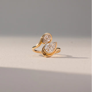 Front view of Toi Et Moi Diamond Ring in 14KT Yellow Gold, featuring pear-shaped and oval lab grown diamonds in fluid wrap settings. Set on a beige backdrop, the design highlights contrast and sculpted elegance.
