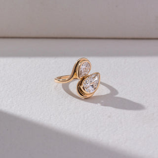 Angled view of Toi Et Moi Diamond Ring in 14KT Yellow Gold, featuring pear-shaped and oval lab grown diamonds in fluid wrap settings. Set on a beige backdrop, the design highlights depth and sculpted elegance.
