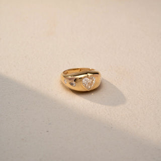 Side view of a heart-cut solitaire signet ring in 14 KT Yellow Gold, featuring a heart-shaped lab-grown diamond with flush-set round diamond accents along the band, displayed against a beige backdrop.