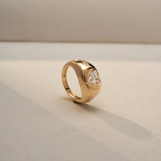 Angled view of a heart-cut solitaire signet ring in 14 KT Yellow Gold, featuring a heart-shaped lab-grown diamond with flush-set round diamond accents along the band, displayed against a beige backdrop.