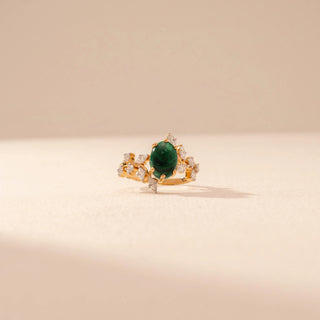 Front view of a 14KT yellow gold ring featuring a green emerald with round and marquise-cut diamonds, set against a beige background.
