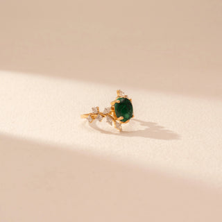 Side view of a 14KT yellow gold ring featuring a green emerald with round and marquise-cut diamonds, set against a beige background.