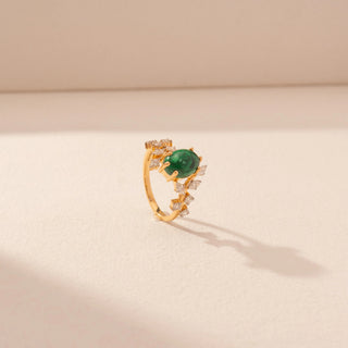 Angled view of a 14KT yellow gold ring featuring a green emerald with round and marquise-cut diamonds, set against a beige background.