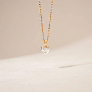 Front view of a heart-shaped diamond pendant in 14KT Yellow Gold, featuring a solitaire stone on a delicate chain against a beige backdrop.