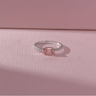 Top view of emerald-cut pink gemstone ring with a pavé diamond band in white gold, set on a soft pink textured surface, highlighting delicate hue and refined, geometric sparkle.
