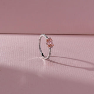 Angled view of emerald-cut pink gemstone ring with a pavé diamond band in white gold, set on a soft pink textured background, highlighting airy design and delicate sparkle.

