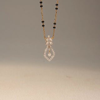 Close up view of a diamond mangalsutra pendant in 14KT Yellow Gold, featuring an open teardrop design with a dangling pear-cut diamond. Captured against a beige backdrop, it exudes graceful sparkle.