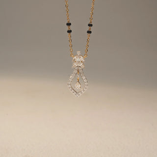 Front view of a diamond mangalsutra pendant in 14KT Yellow Gold, featuring an open teardrop design with a dangling pear-cut diamond. Captured against a beige backdrop, it exudes graceful sparkle.