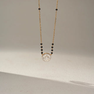Close-up view of a diamond halo mangalsutra necklace in 14 KT Dual Gold, featuring an oval-cut lab-grown diamond encircled by a sparkling halo, suspended from a delicate chain with black bead accents against a beige backdrop.
