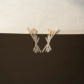 Angle  view of diamond statement earrings in 14 KT Yellow Gold, featuring intersecting pavé-set bars arranged in an abstract burst design against a neutral textured backdrop.
