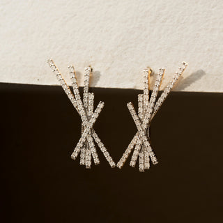 Close view of diamond statement earrings in 14 KT Yellow Gold, featuring intersecting pavé-set bars arranged in an abstract burst design against a neutral textured backdrop.
