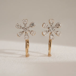 Floral diamond hook earrings in 14KT Yellow Gold, designed with six sparkling petals and a central round diamond, are displayed upright on a light surface with soft natural lighting.