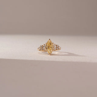 Front view of a marquise-cut yellow lab-grown diamond ring in yellow gold, featuring tapered side diamonds. Captured against a beige backdrop, the elegant cluster design highlights radiant brilliance.