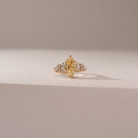 Front view of a marquise-cut yellow lab-grown diamond ring in yellow gold, featuring tapered side diamonds. Captured against a beige backdrop, the elegant cluster design highlights radiant brilliance.