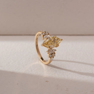 Angled side view of a yellow gold ring featuring a marquise-cut yellow lab-grown diamond centre stone, accented by marquise and round side diamonds. Captured against a beige backdrop, the elevated setting highlights brilliance and dimension.