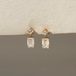 Front view of diamond drop earrings in yellow gold, featuring princess-cut diamond studs paired with dangling emerald-cut diamonds. Displayed against a neutral backdrop, the design exudes modern elegance.