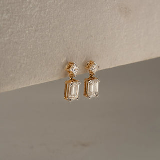 Side view of diamond drop earrings in yellow gold, featuring princess-cut diamond studs paired with dangling emerald-cut diamonds. Displayed against a neutral backdrop, the profile highlights refined craftsmanship.