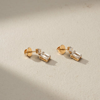 Angled view of diamond drop earrings in yellow gold, featuring princess-cut diamond studs paired with dangling emerald-cut diamonds. Set against a beige backdrop, the design showcases elegant brilliance.