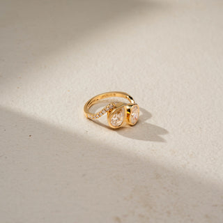Side view of a bypass diamond ring in 14 KT Yellow Gold, featuring bezel-set pear-shaped and oval lab-grown diamonds with pavé diamond accents along the split band, displayed against a beige backdrop.